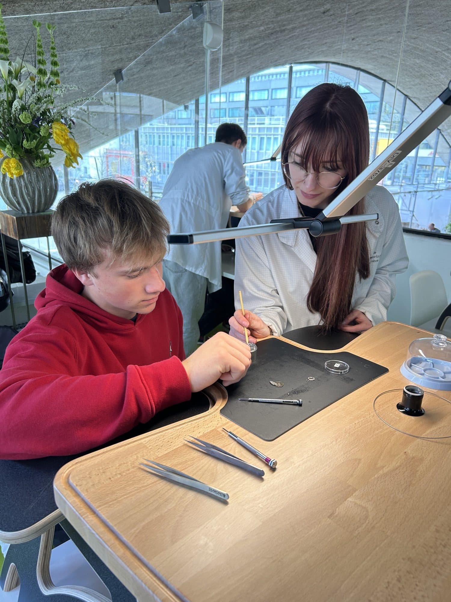 Un adolescent s'initie à l'horlogerie sur un établi, pinces et loupe à portée de main, accompagné d'une jeune femme en blouse blanche